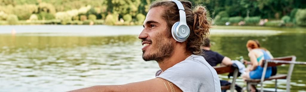 Mann sitzt lächelnd und mit Kopfhörern an einem See.