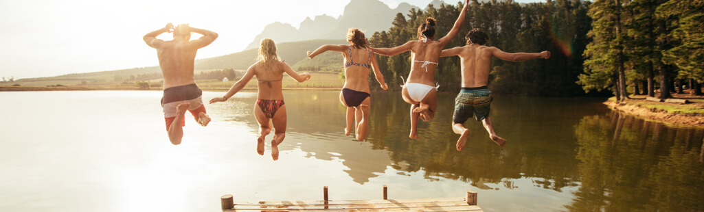 Eine Gruppe von 5 Personen in Badekleidung spring von einem Steg gleichzeitig in einen See. Das Licht ist warm und es sieht sommerlich aus.
