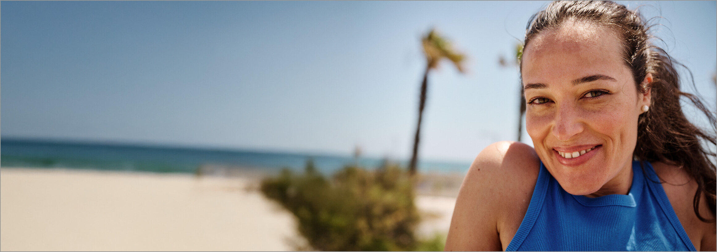 Eine sommerlich gekleidete Frau schaut lächelnd in die Kamera. Im Hintergrund sind Strand, Palmen und das Meer zu sehen. 