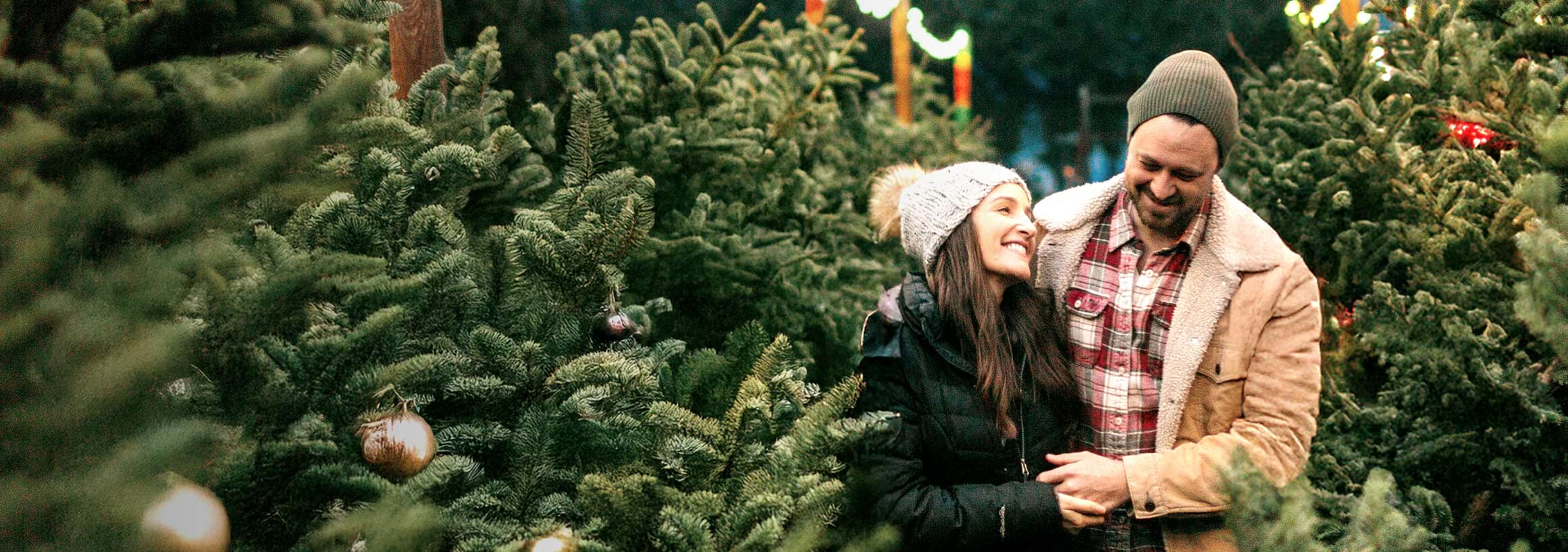 Ein winterlich gekleidetes  Pärchen steht fröhlich zwischen Tannenbäumen.